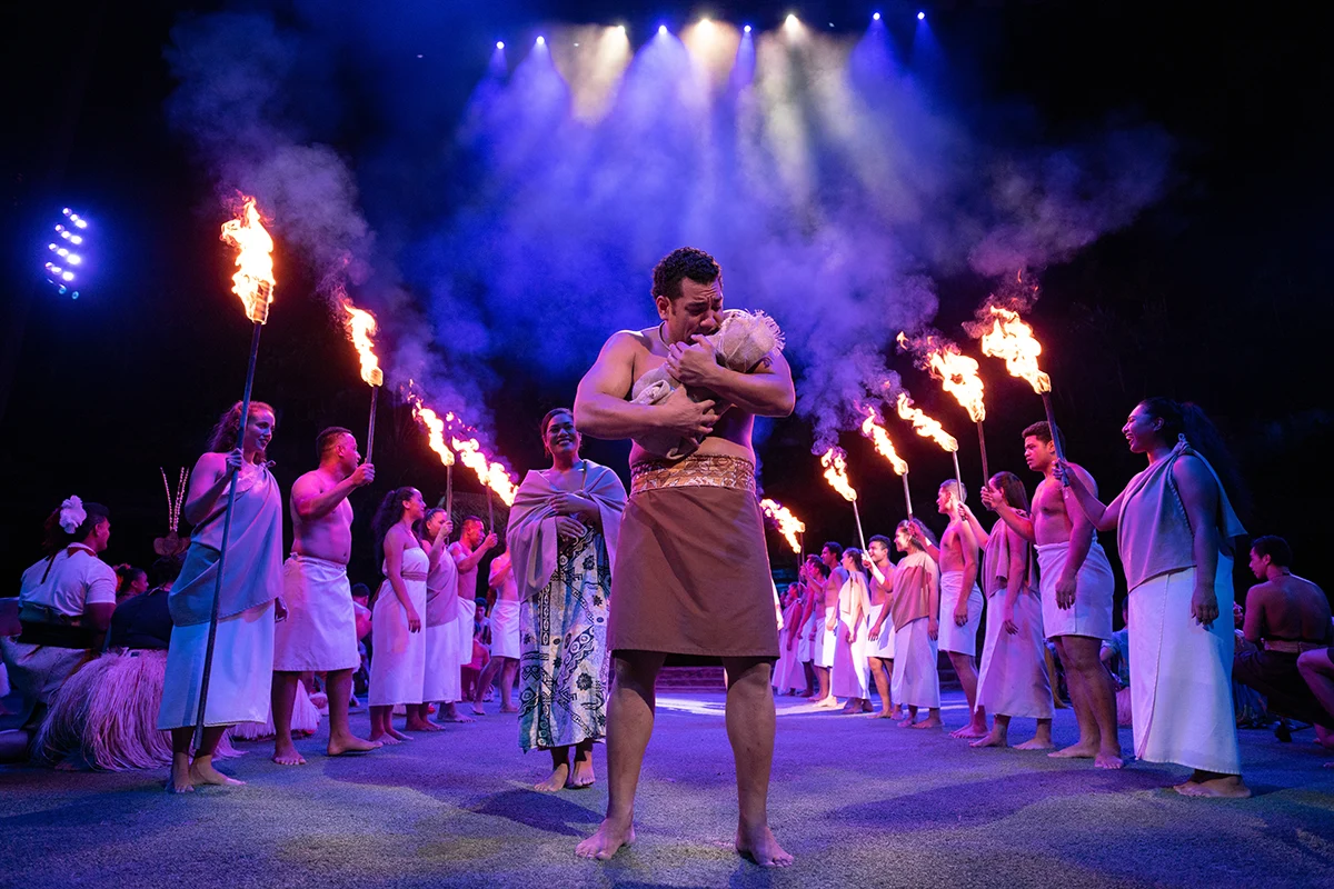 A performer cradles a newborn at the center of a ceremonial stage, surrounded by torch-bearers in traditional Pacific Islander dress under dramatic blue stage lighting and smoke