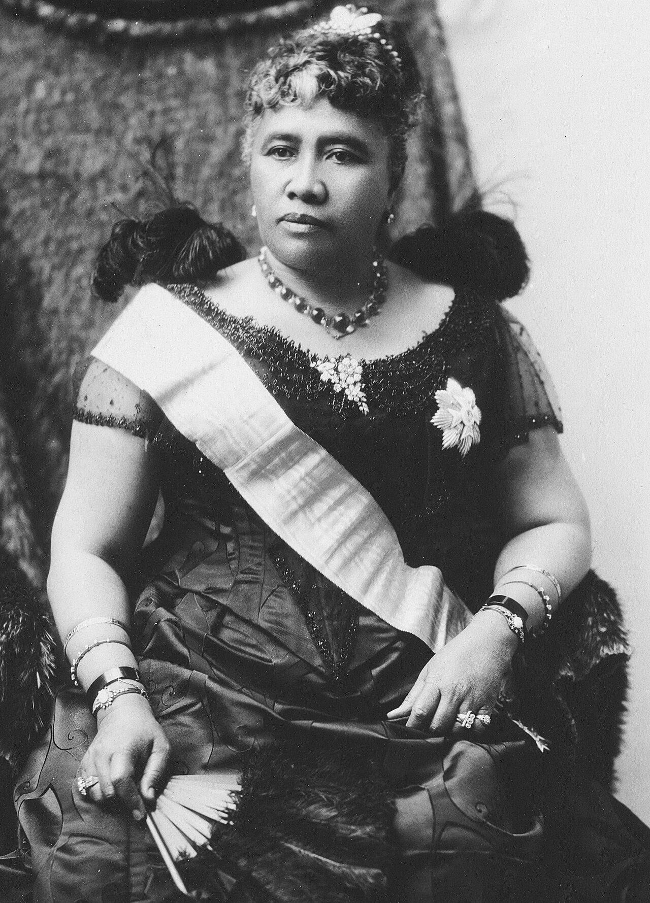 Queen Liliʻuokalani, seated, wearing a royal sash and holding a fan, c. 1891